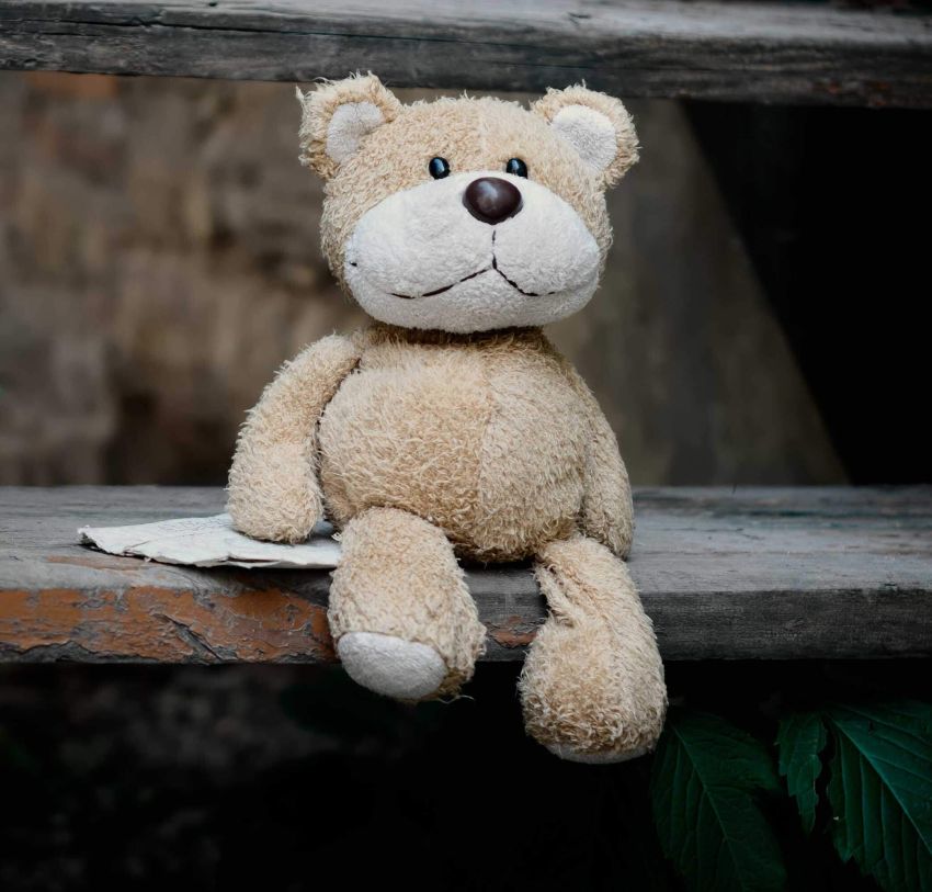 Das Bild zeigt einen Teddybär auf einem Holzbrett sitzend und einem Blatt Papier