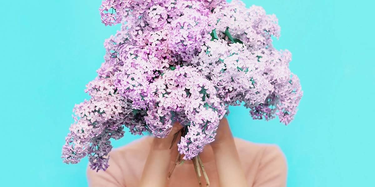 The picture shows a large lilac bouquet. A woman is hiding behind the bouquet. The picture stands for the pause we should take regularly to lead a healthy life.