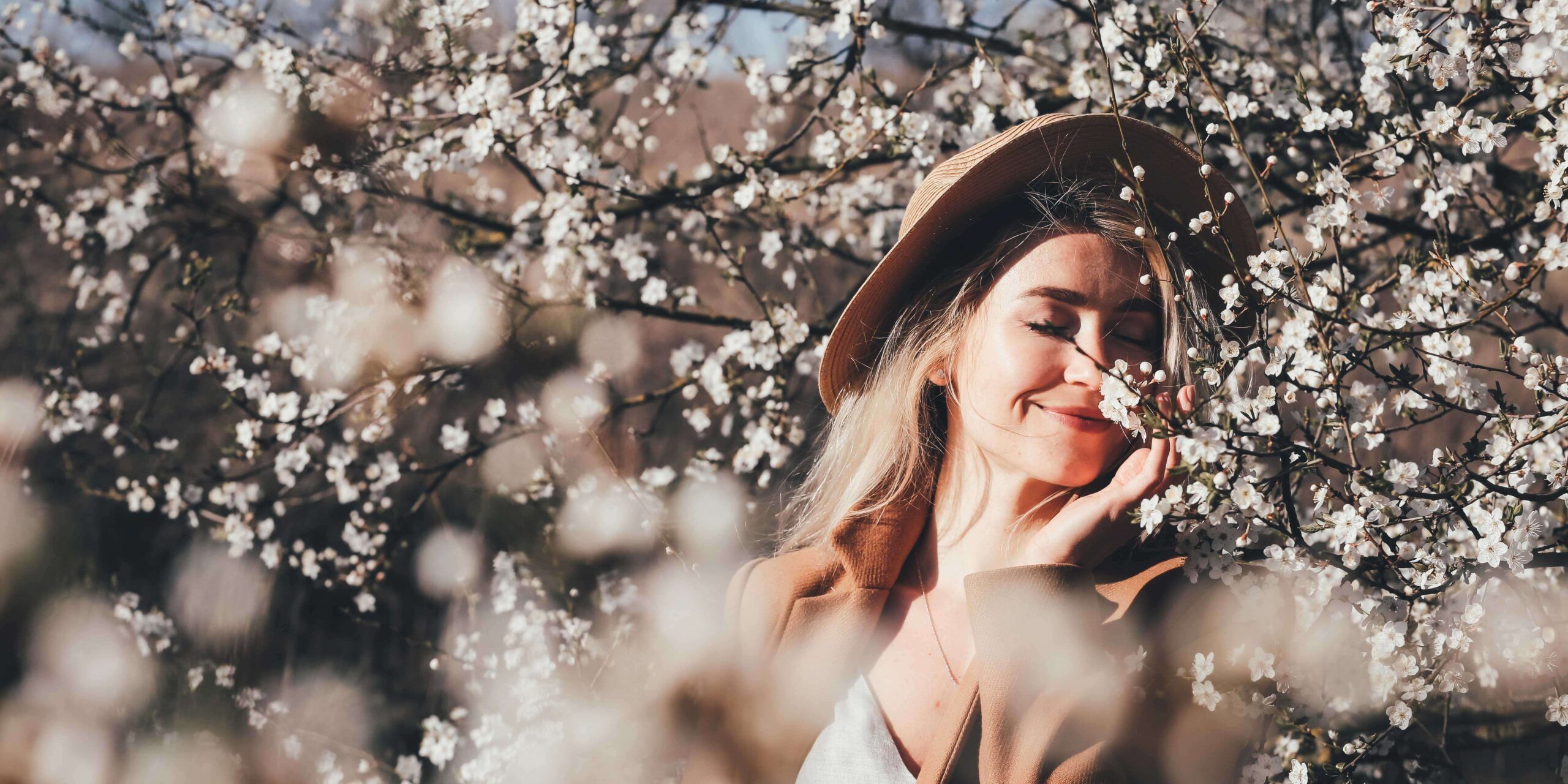 The image shows a young woman with blond, long hair and wearing a hat. She sniffs on the blossoms of a tree.