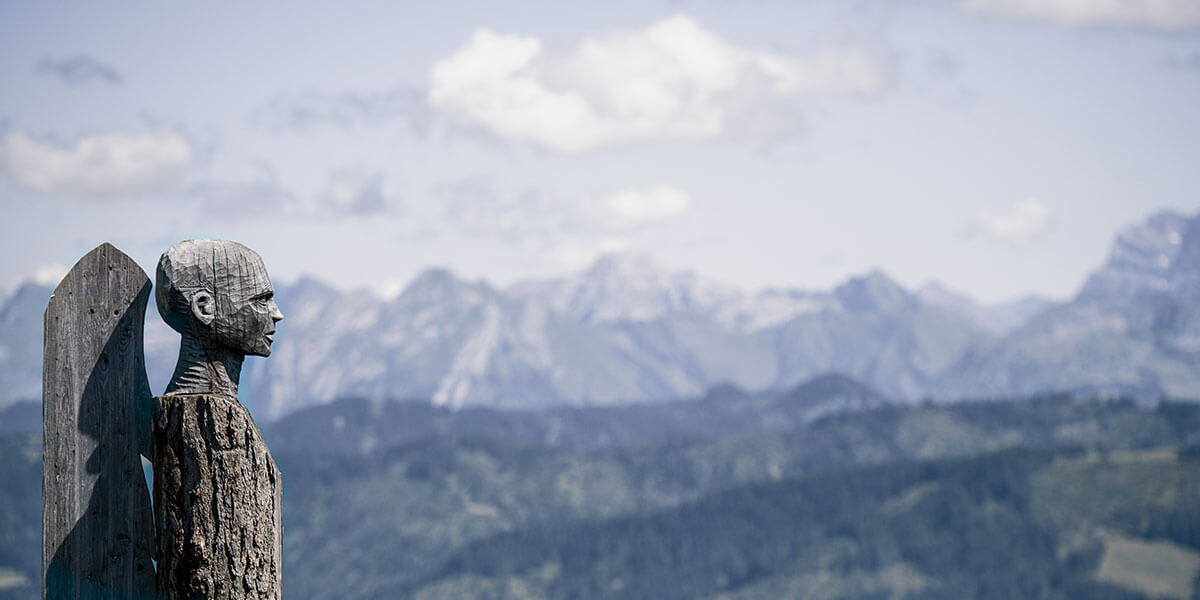 The picture shows a carved wooden figure with wings standing on a mountain with a mountain panorama in the background. It stands for the fitness of the lungs and what we can do for it.